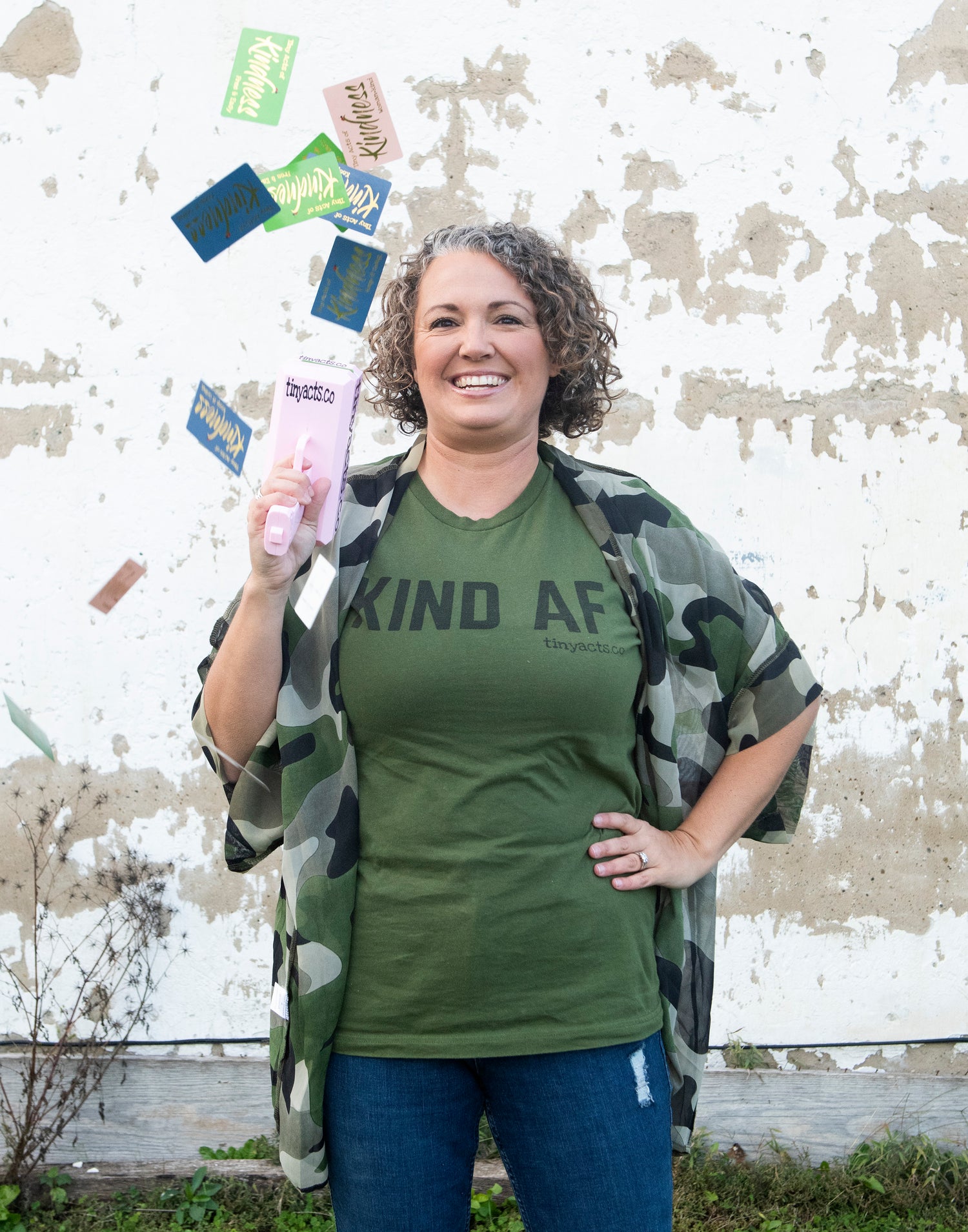 Gina Sachs, Kind AF, camo, kindness cards, tinyacts.co, female business owner, money gun, curly hair, army green, small business owner, women owned business, business headshot, hand on hip, kind as fuck